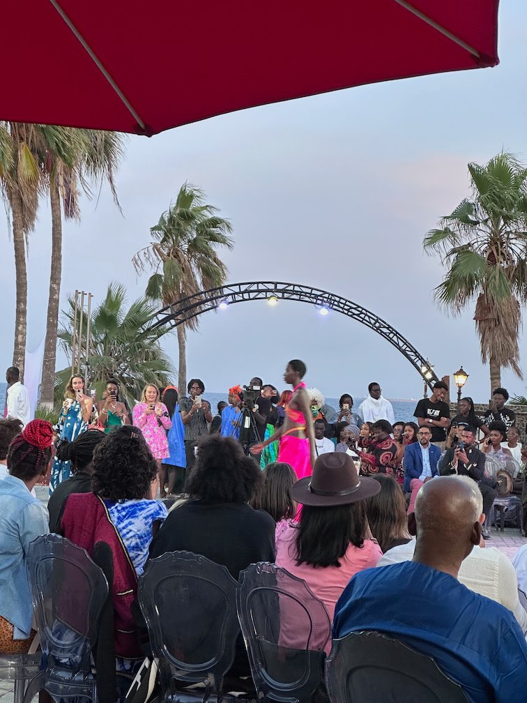 People watch a fashion show under palm trees at sunset on Goree Island.