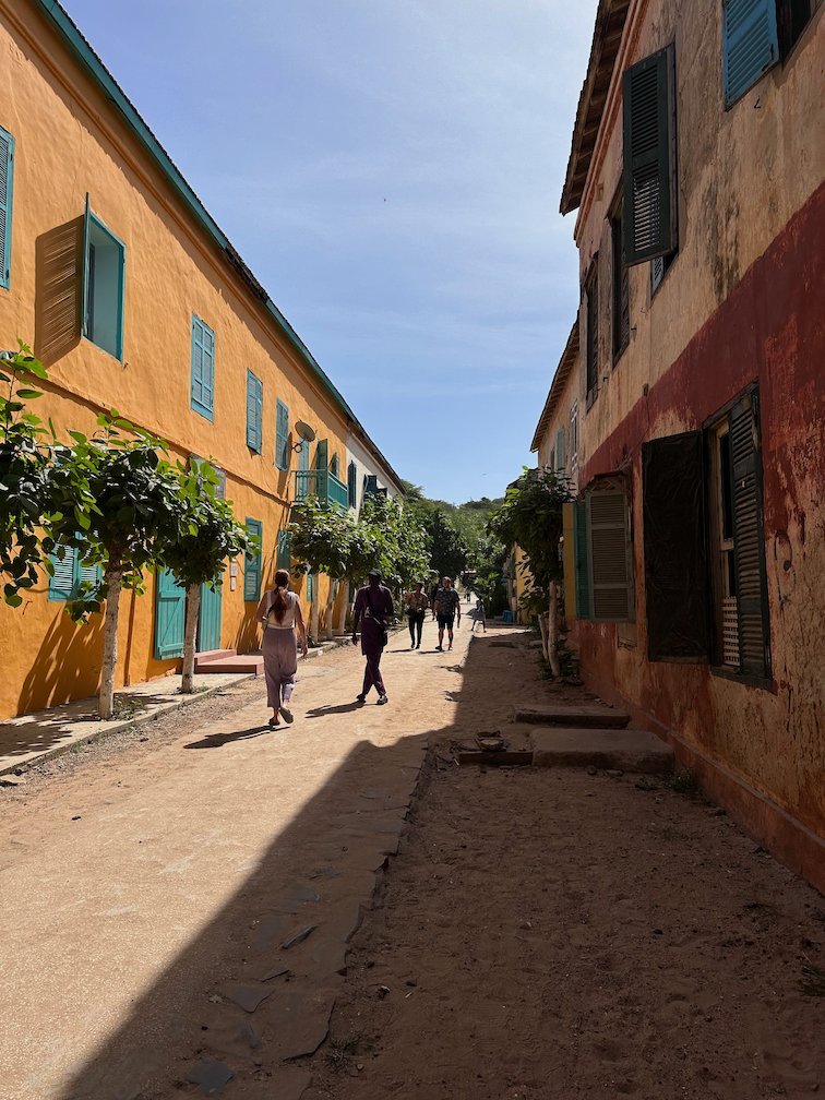 People walk on a sandy street between orange houses on Goree Island.