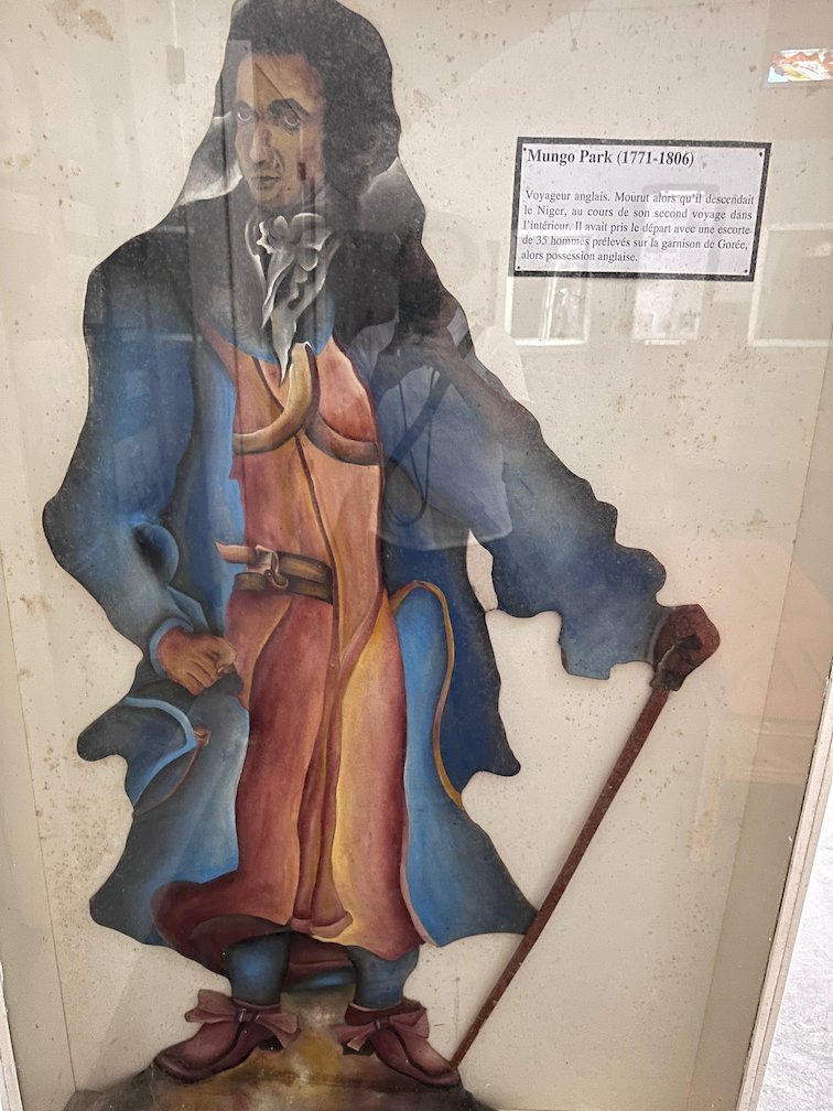 A painting shows a man in a long blue coat with a stick on Goree Island.