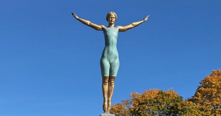 Bronze statue of diver Greta Johansson with her arms spread wide against a blue sky and autumn trees.