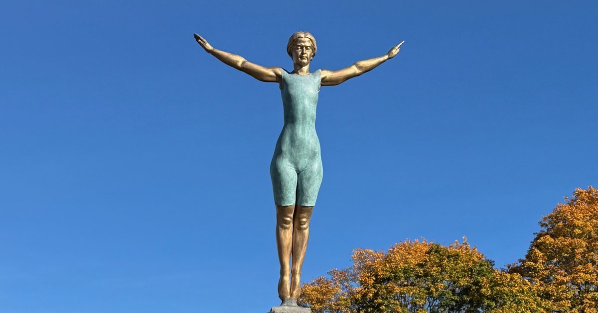 Bronze statue of diver Greta Johansson with her arms spread wide against a blue sky and autumn trees.