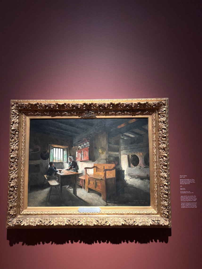 Painting by Harriet Backer of a woman at a desk by a window, on a red museum wall
