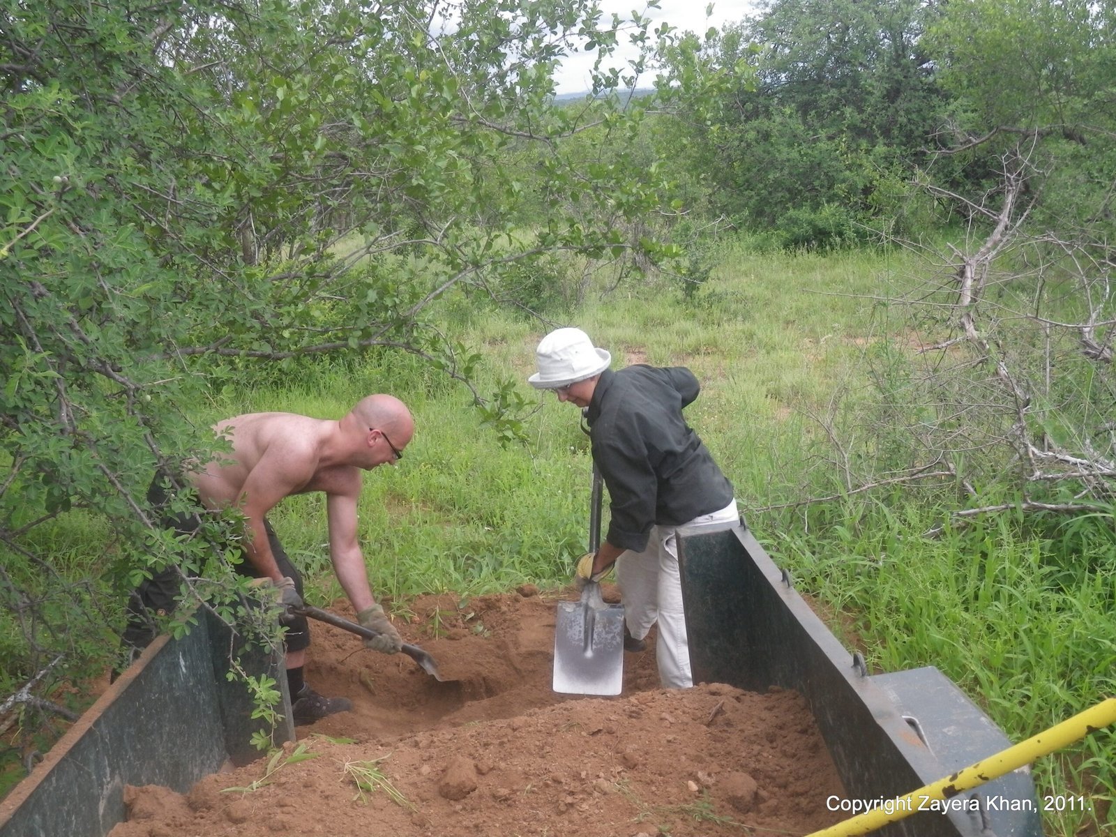 Two people shovel soil from a trailer on a bush road near Hoedspruit, South Africa.