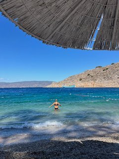 Swimmer in clear blue water seen from under a straw umbrella on a beach in Hydra