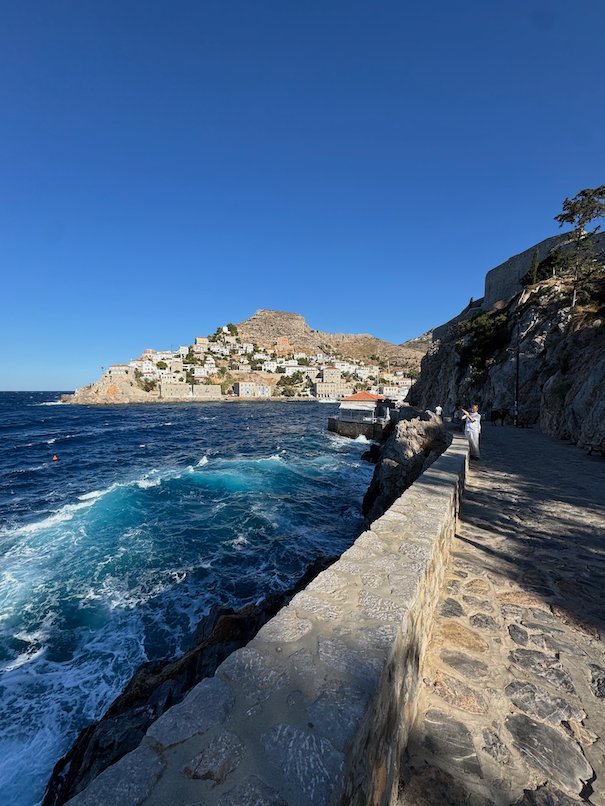 Stone path along the coast with waves and Hydra town ahead