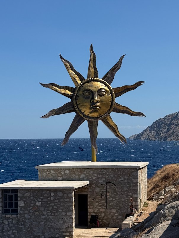 Golden sun sculpture with a face on a stone building by the sea in Hydra