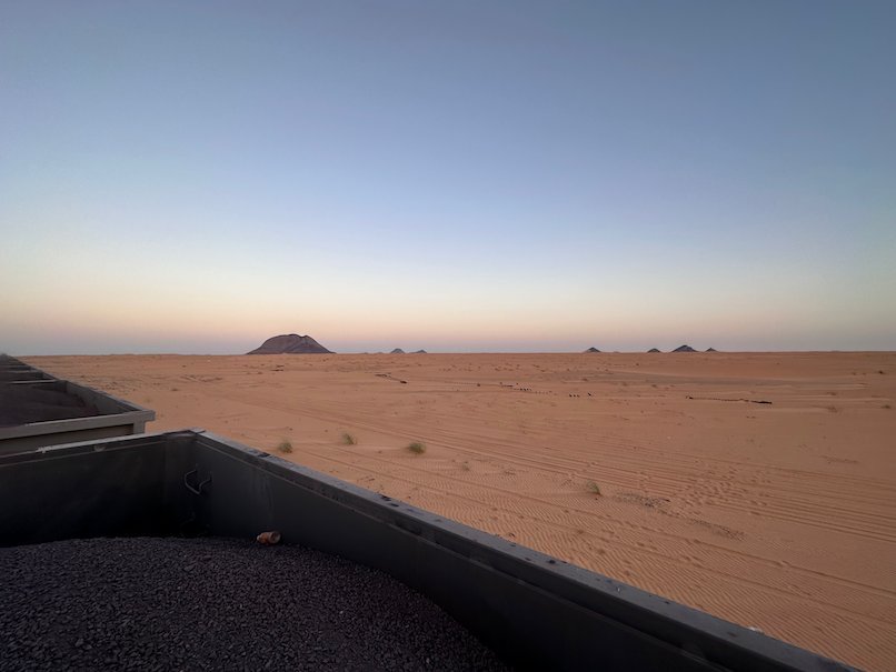 An ore wagon passes flat desert with a lone rock hill in the morning.
