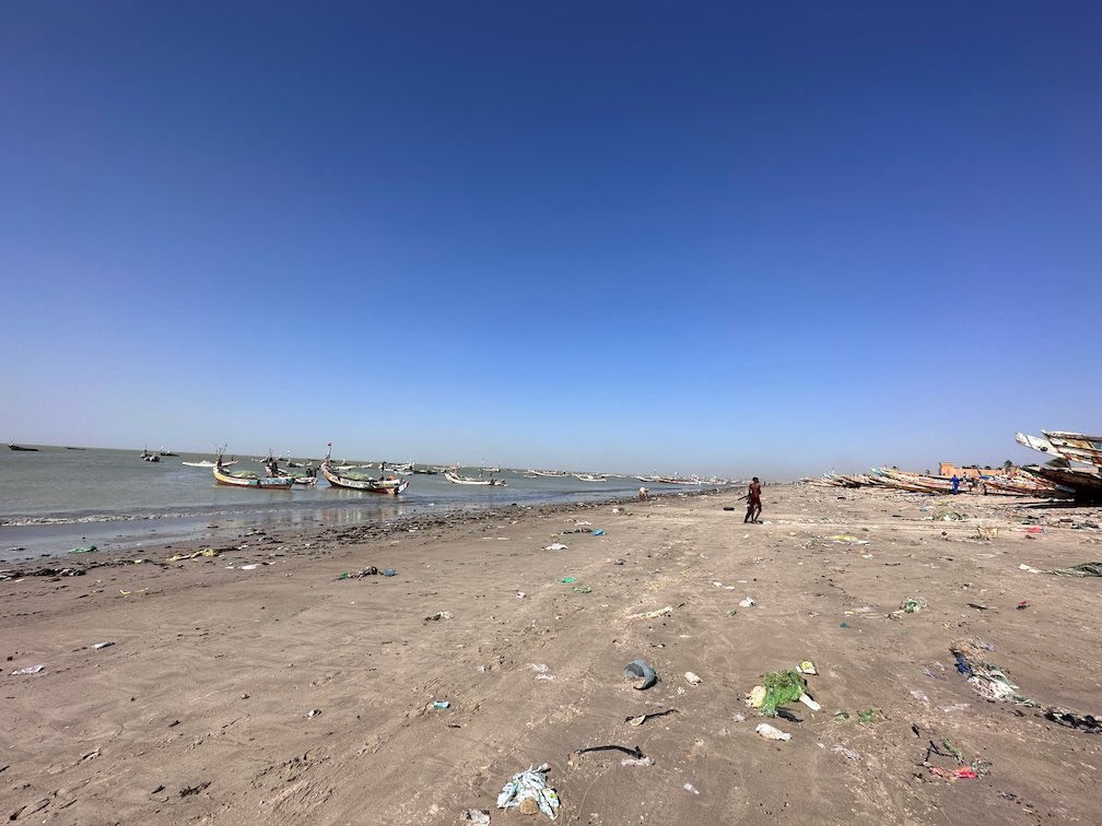 Boats lie off a beach with litter on the sand in Joal.