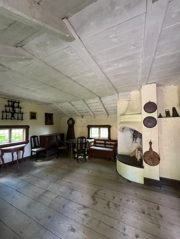 Large farmhouse room with white walls, a round tiled stove and old furniture