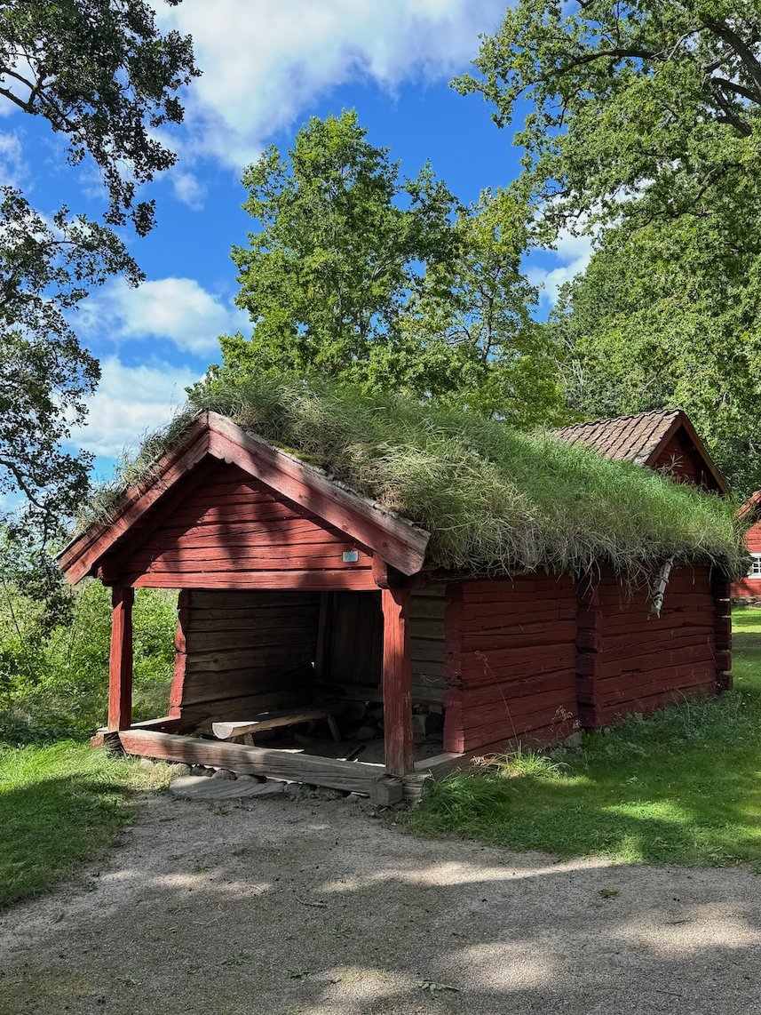 Red wooden cottage with a grass roof and an open front, under green trees