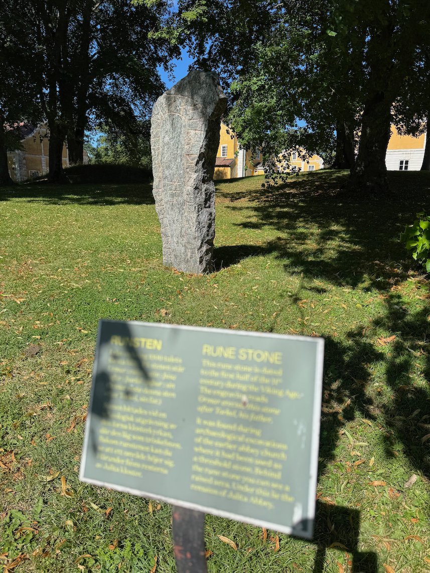 Tall rune stone in the grass under trees at Julita Gård, with an information sign in front