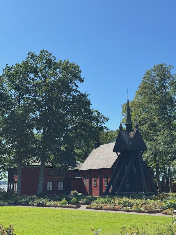 Small dark wooden chapel with a spire beside red wooden buildings and large trees