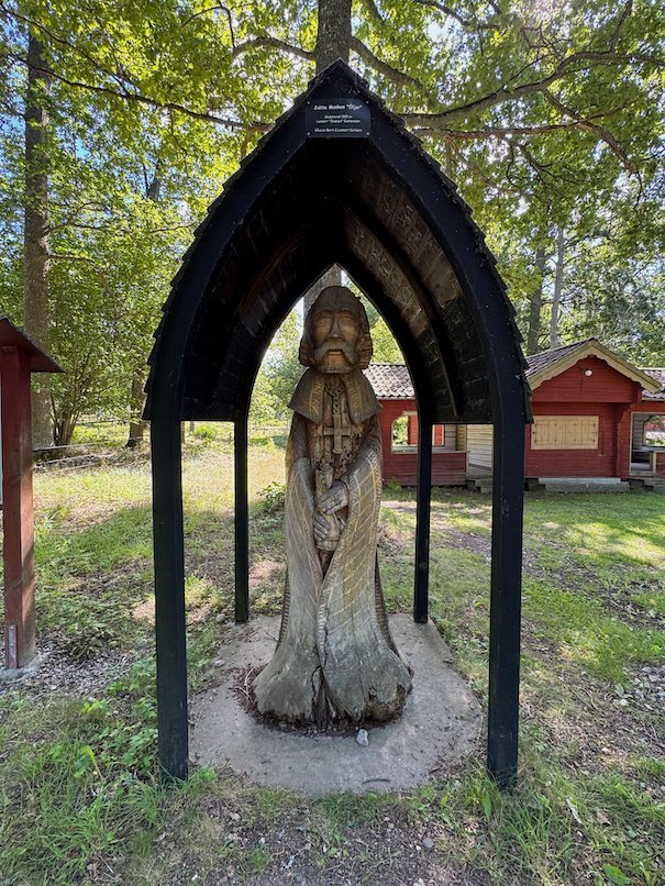 Carved wooden monk sculpture under a black wooden shelter, with red buildings behind