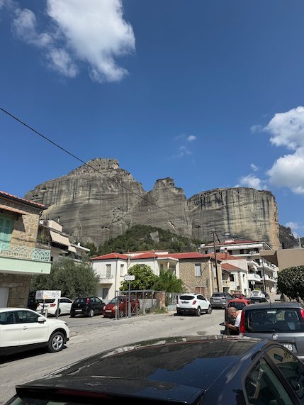 Houses and parked cars on a street below the huge rocks of Meteora