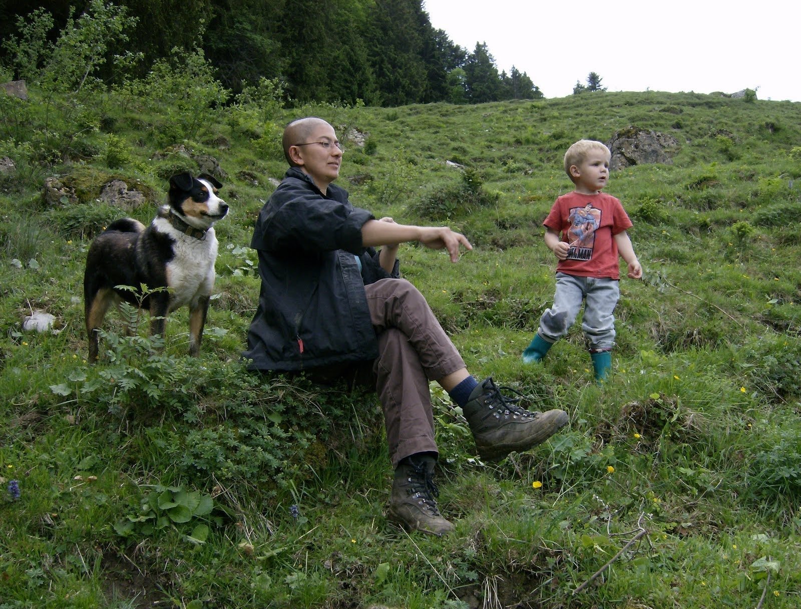 A person and a dog sit on a grassy slope while a child plays nearby.
