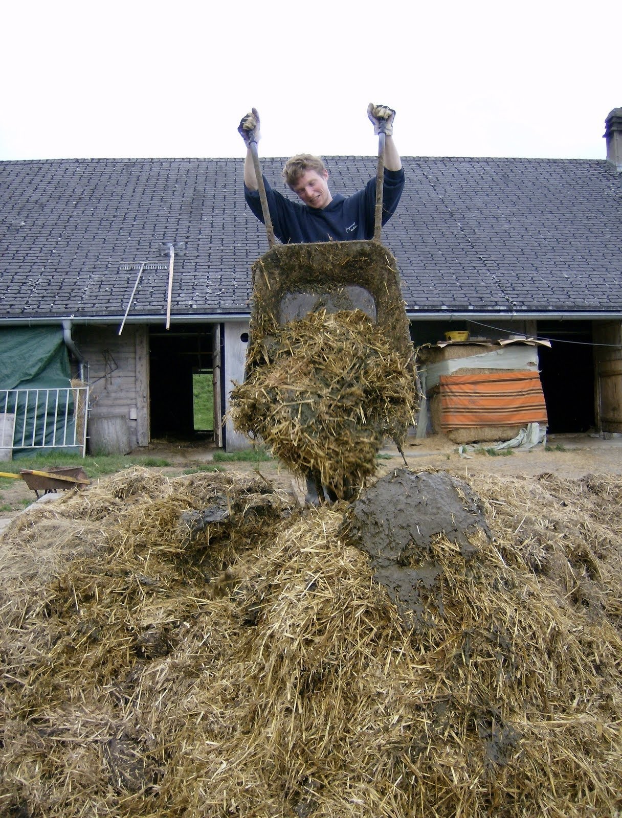 A young man lifts a wheelbarrow over a manure heap in front of a barn.