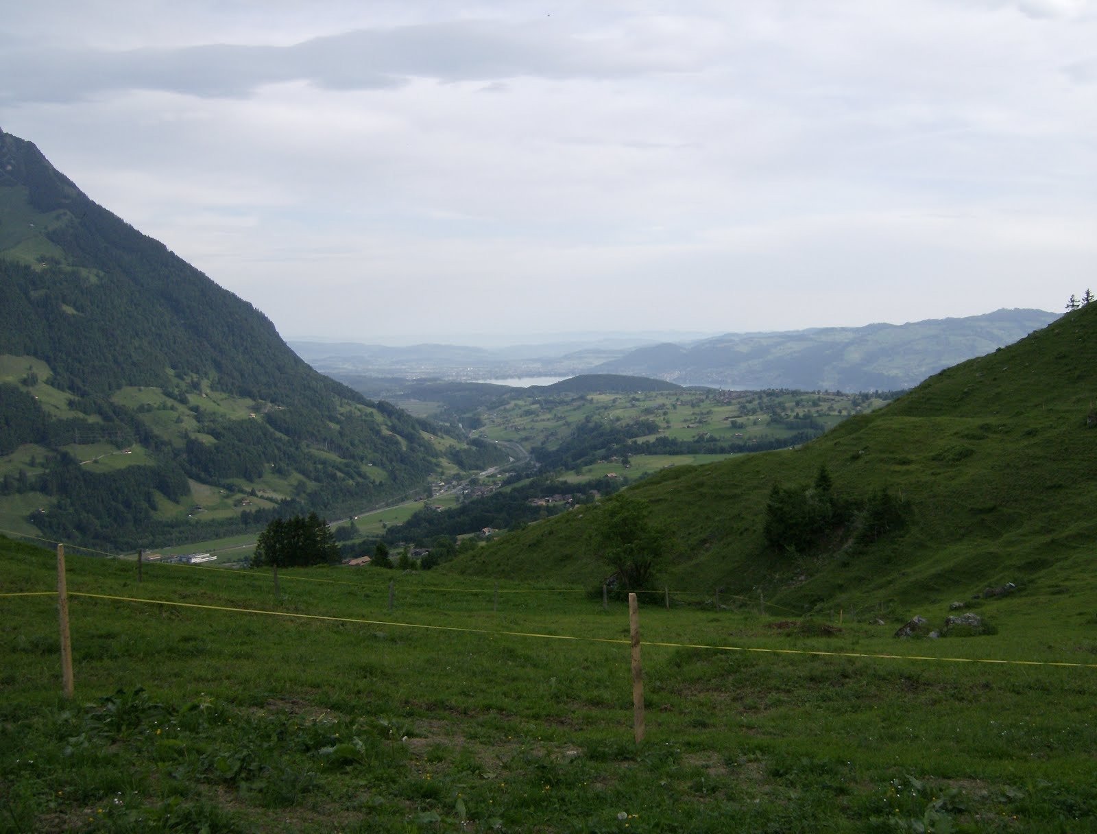 A fenced pasture looks down a green valley toward a distant lake in Switzerland.