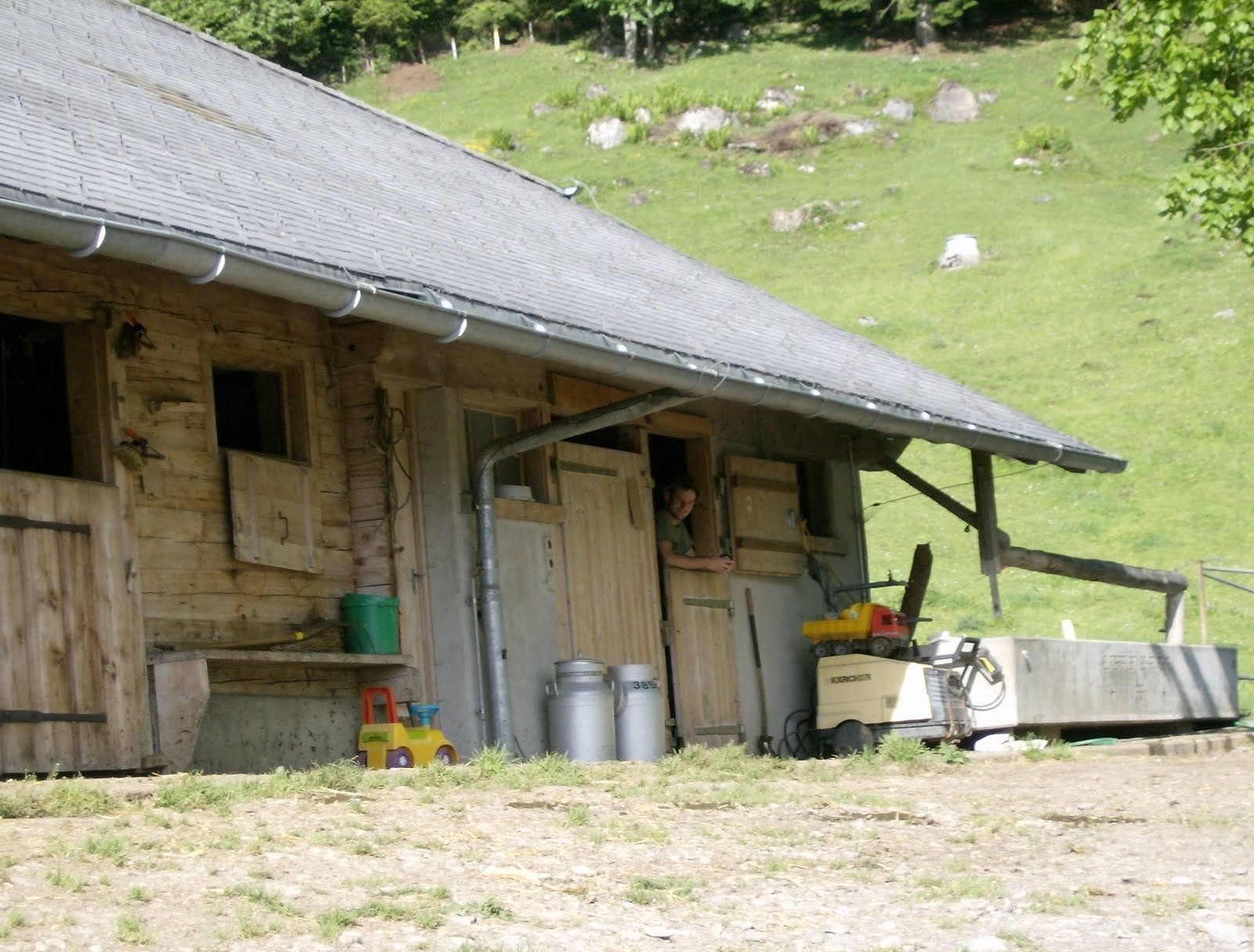 A wooden alp farmhouse with milk cans stands below a green slope in Switzerland.