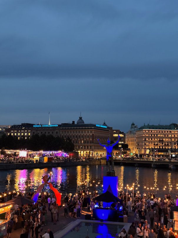Crowds on the quay by Strömmen at dusk, with lit buildings and festival lights reflected in the water