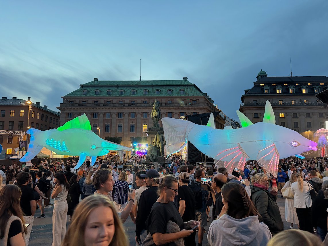 Large glowing fish lanterns carried above the crowd on a square in central Stockholm at dusk