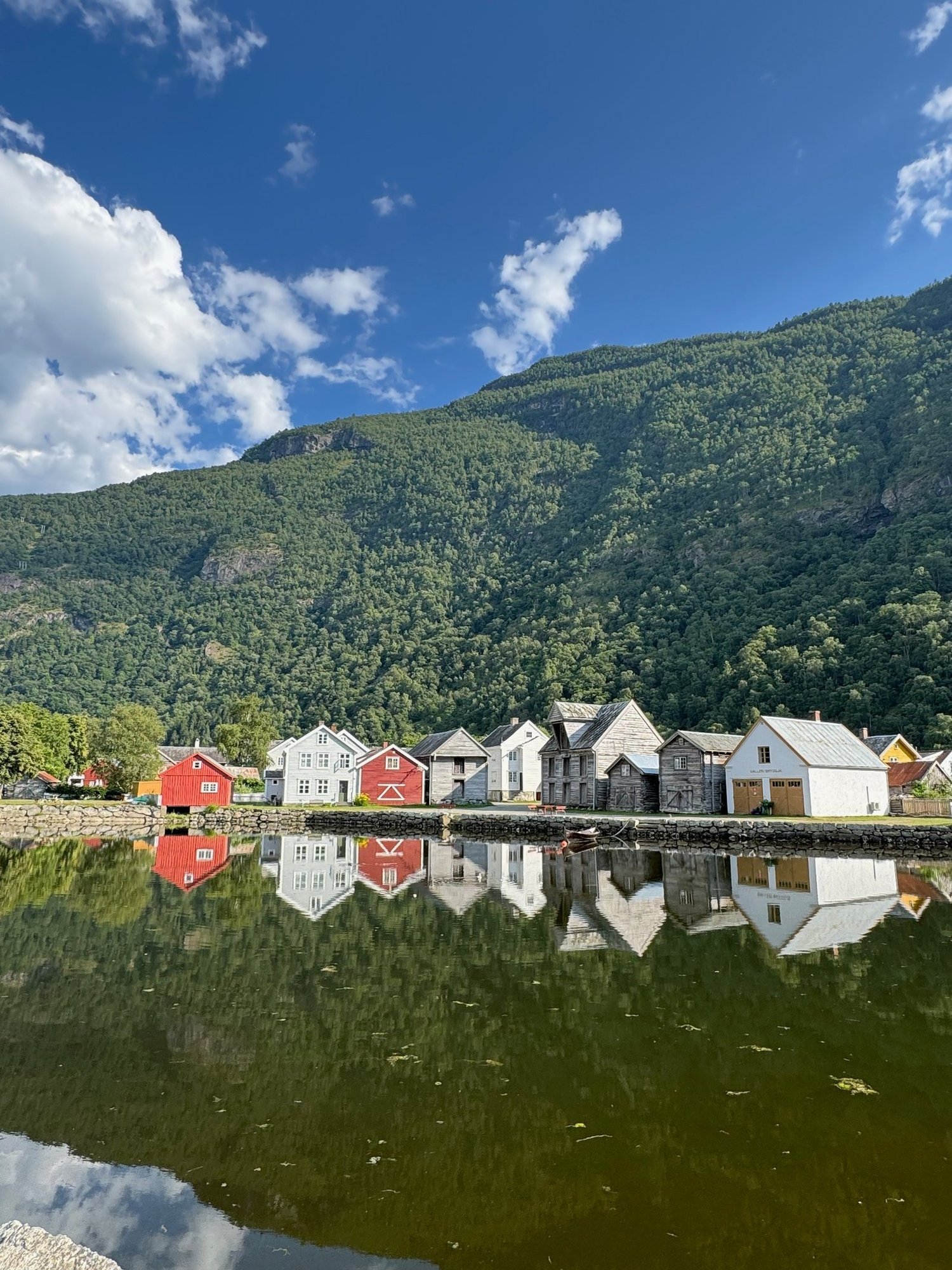 Red and white boathouses reflected in calm water at Lærdalsøyri, with green mountains behind