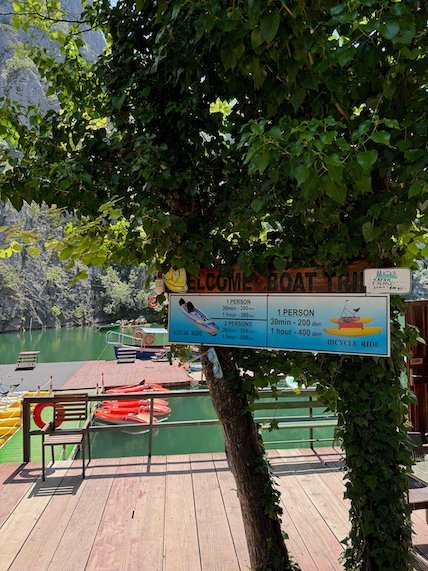 A boat trip sign hangs from a tree above a wooden dock in Matka Canyon.