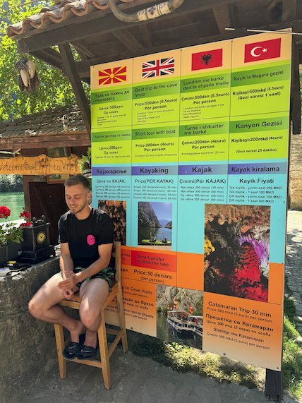 A man sits next to a board with boat and kayak prices in four languages at Matka Canyon.