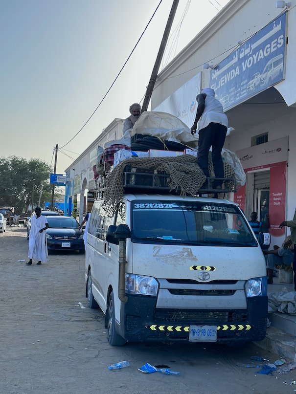 Men load luggage on the roof of a white minibus in a Mauritanian town.