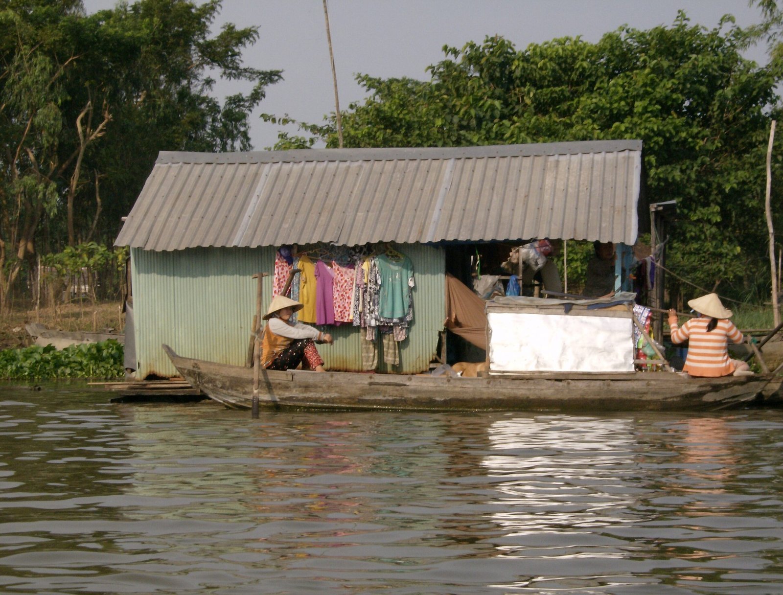 People sit outside a small floating house with a tin roof on the river in the Mekong Delta.