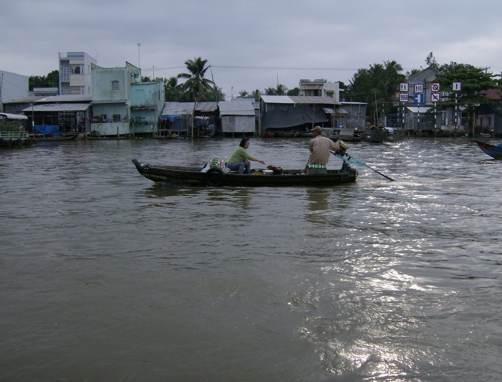 Two people row a long wooden boat across a wide river in the Mekong Delta.
