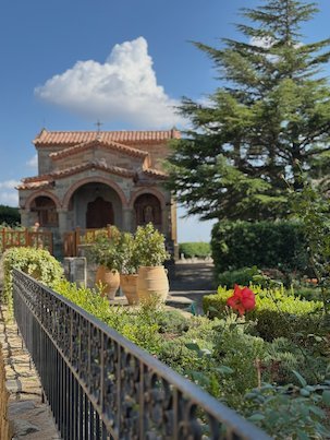 Small stone chapel in a garden with flowers and a cypress tree at Meteora
