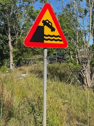 Red warning sign that shows a car falling off a quay into water, beside a path on Möja