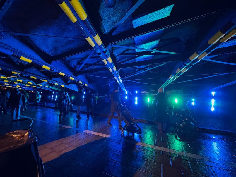 People walking through an underpass with steel beams lit in blue and green light