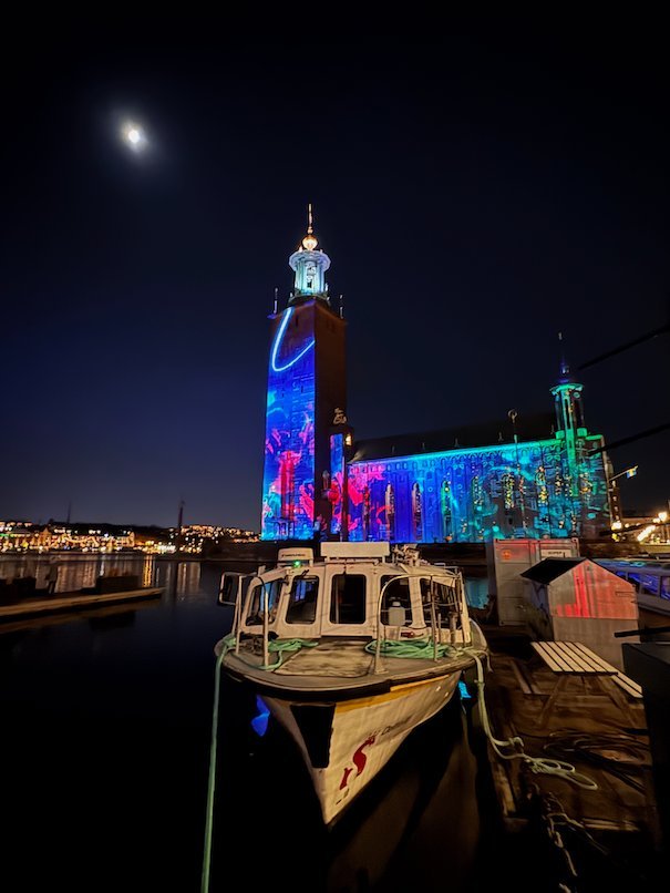 Stockholm City Hall with a blue and pink light projection, a small wooden boat moored in front
