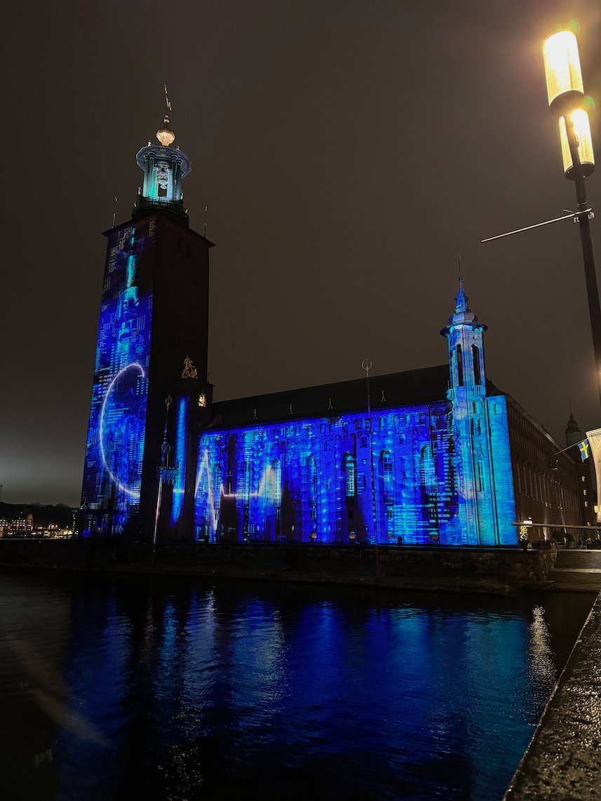 Stockholm City Hall covered in a blue light projection on a cloudy night, reflected in the water