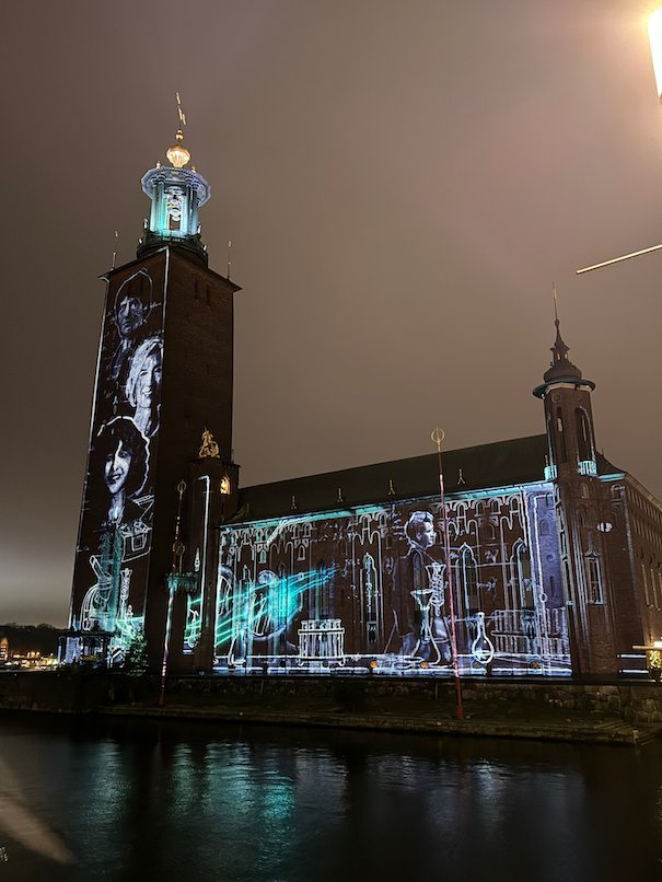 Cyan line drawings of a woman and laboratory scenes projected on Stockholm City Hall on a cloudy night
