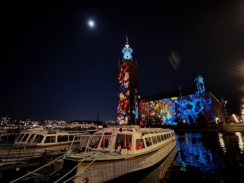 Stockholm City Hall with a red and blue light projection, seen behind tour boats at a jetty under the moon