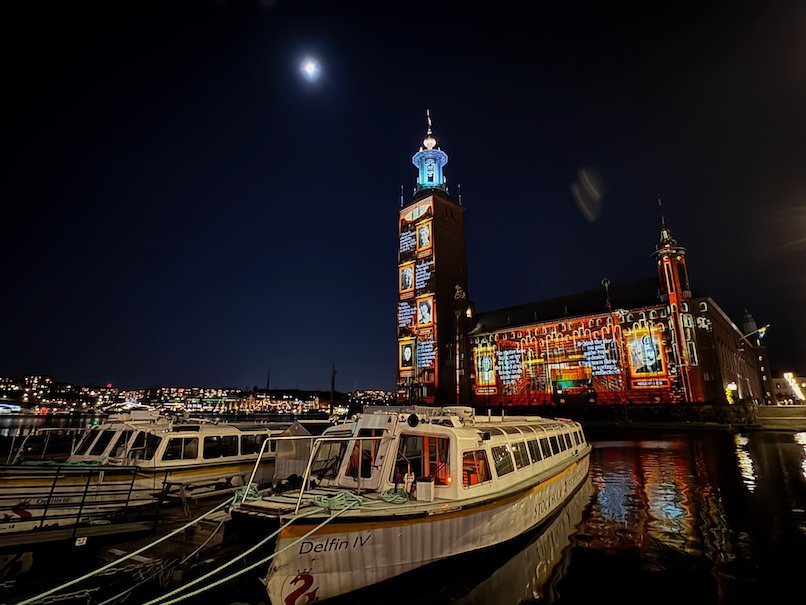 Stockholm City Hall with orange text panels projected on the tower and facade, tour boats in front
