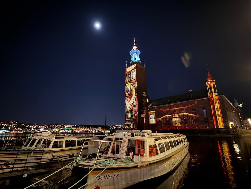 Stockholm City Hall with a red and gold projection on the tower, seen across the water from a jetty with tour boats