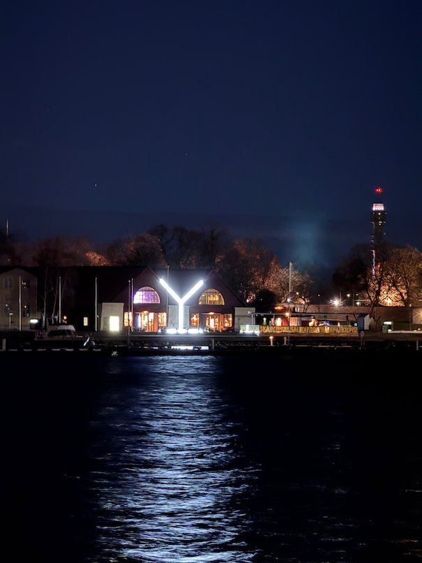 Large illuminated letter Y by the water at night, with its light reflected on the surface