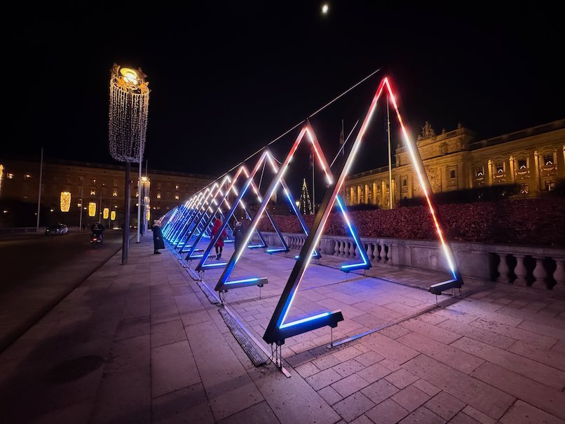 Row of neon triangle light frames on a stone terrace at night, with a lit palace facade behind