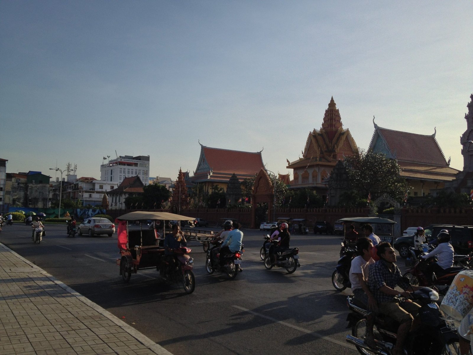 Tuk-tuks and motorbikes pass Buddhist temple roofs on a street in Phnom Penh.