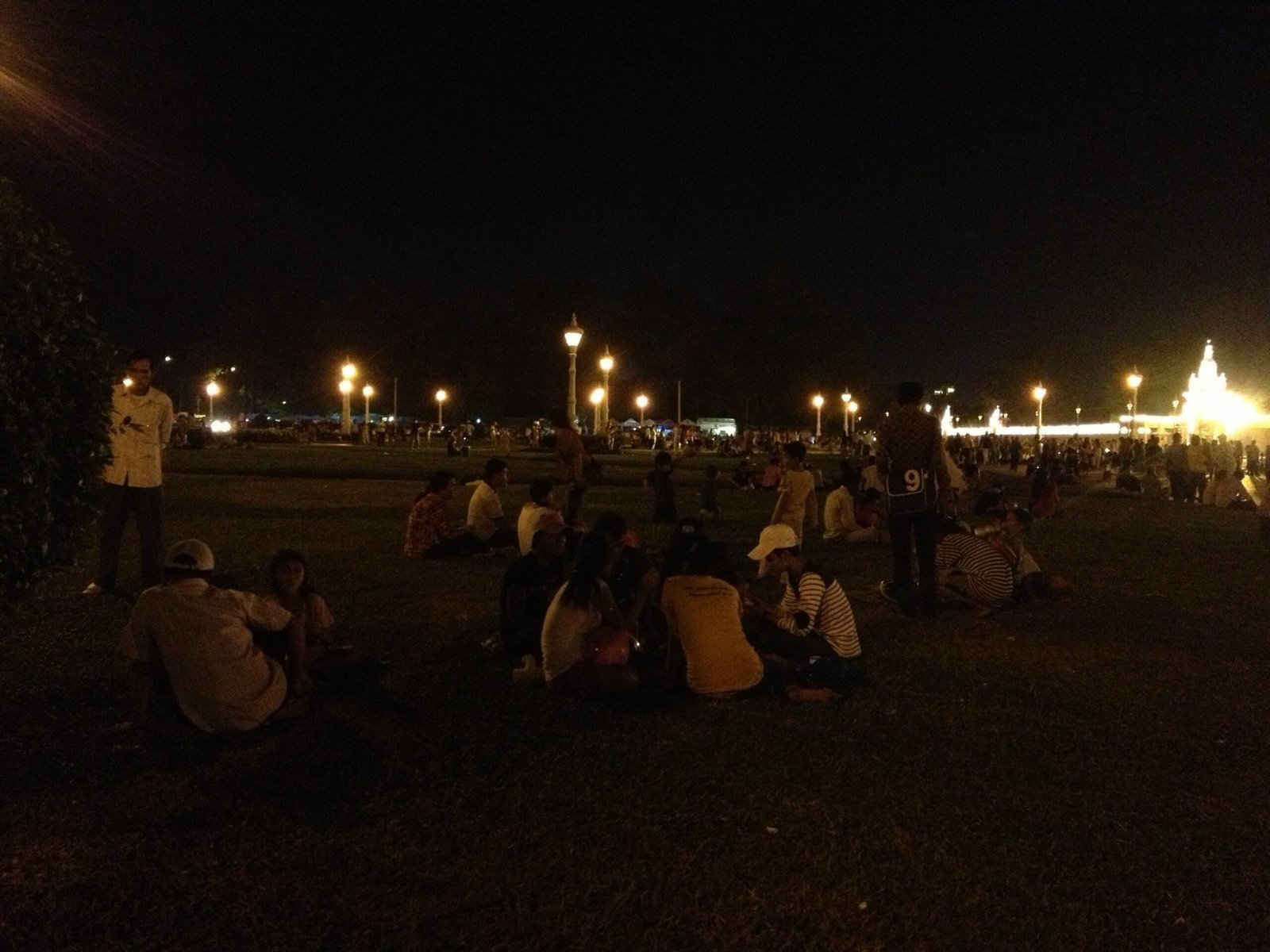 Families sit on the grass of a large square in Phnom Penh at night.