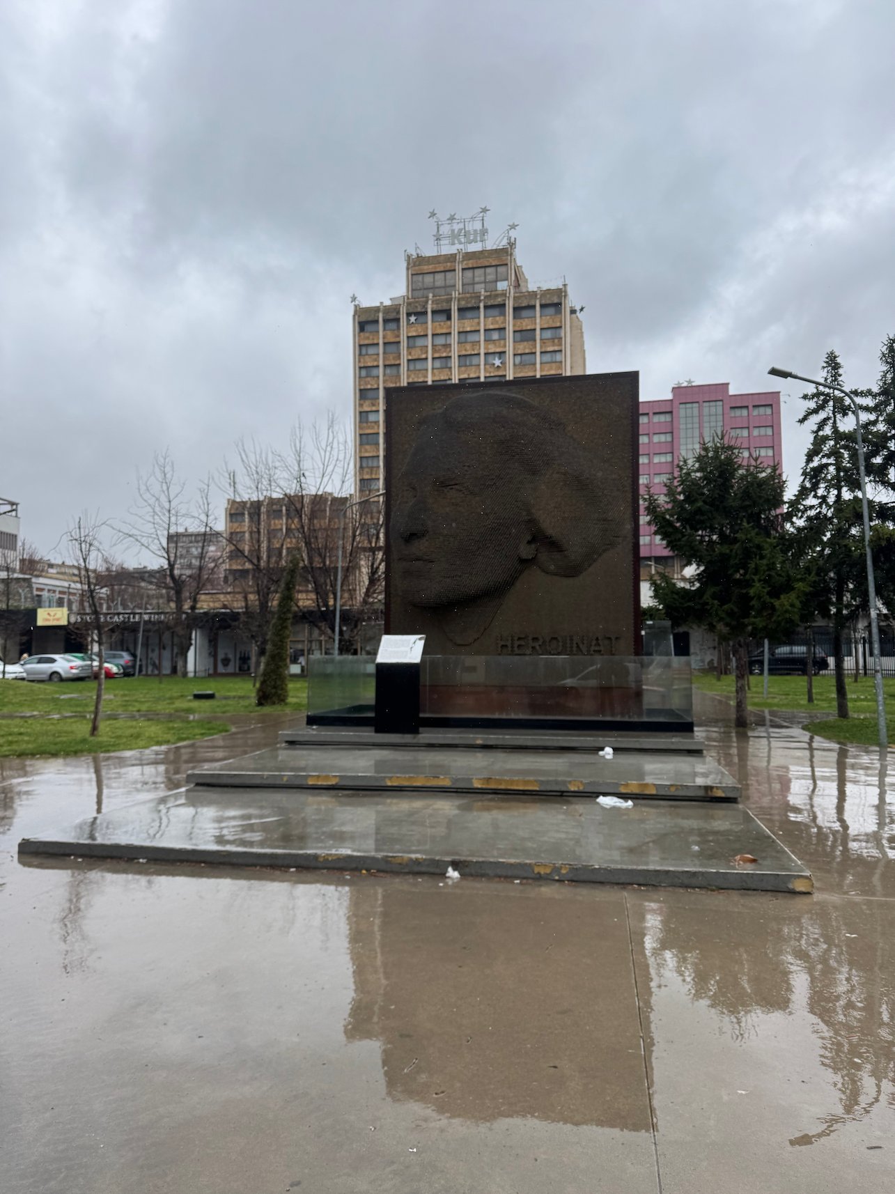 The Heroinat memorial stands in a wet park with tall buildings behind it in Pristina.