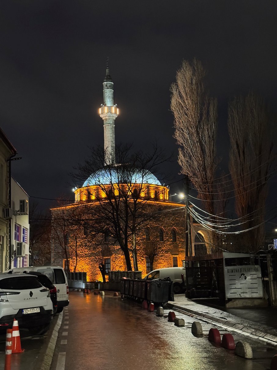 A mosque with a lit dome and a tall minaret stands on a wet street at night in Pristina.