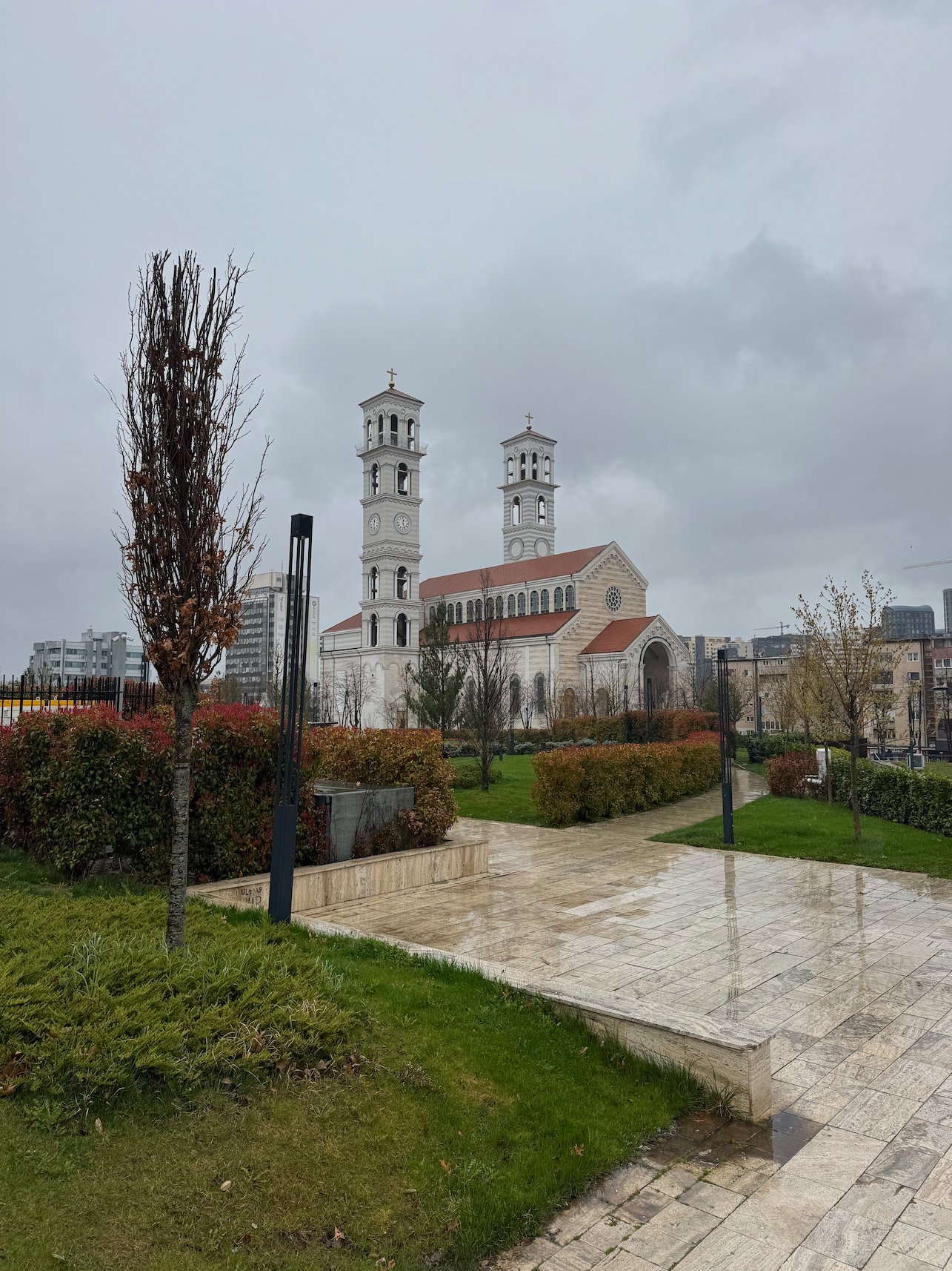 The Cathedral of Saint Mother Teresa with two bell towers stands beyond a wet park in Pristina.