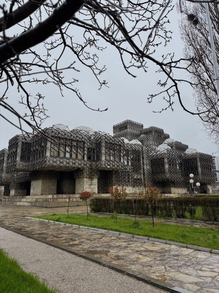 The National Library with white domes and a metal grid facade stands behind bare branches in Pristina.