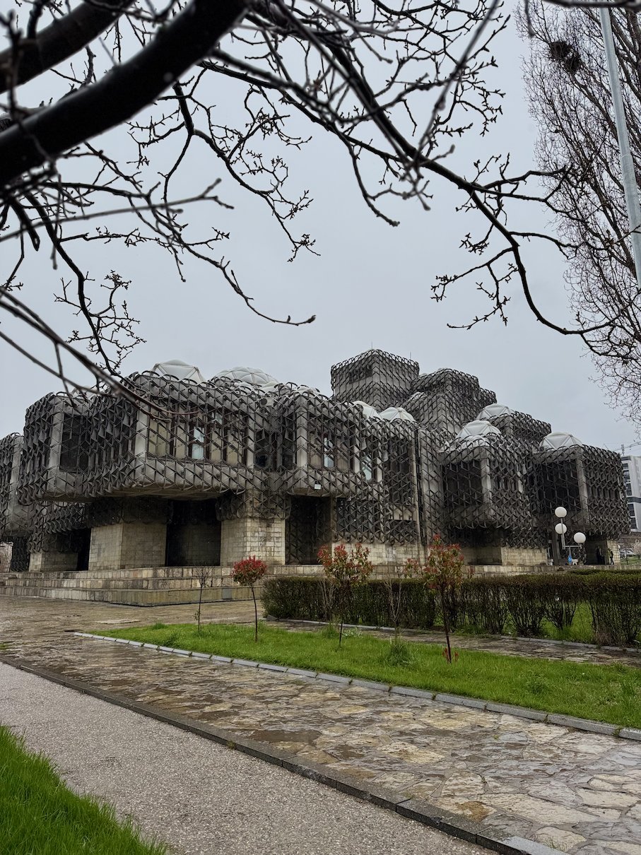 The National Library with white domes and a metal grid facade stands behind bare branches in Pristina.