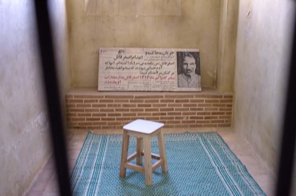 Prison cell with a wooden stool, a green carpet and a plaque at the Qasr Prison Museum