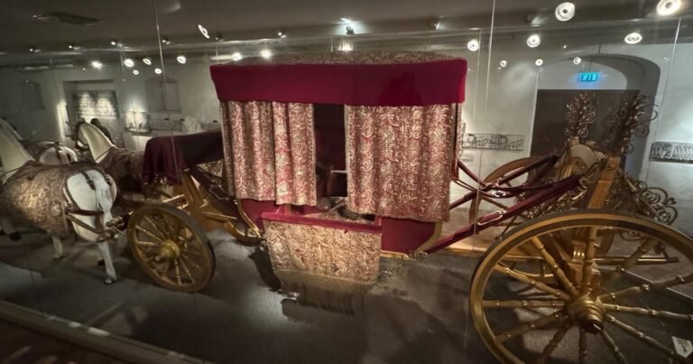 Queen Kristina’s coronation coach from 1650 behind glass, with gilded wheels, red and gold embroidered curtains and white model horses.